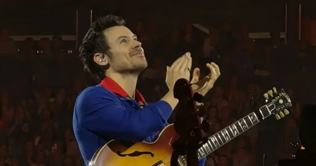 Harry Styles no palco, com um violão e roupa azul e vermelha.