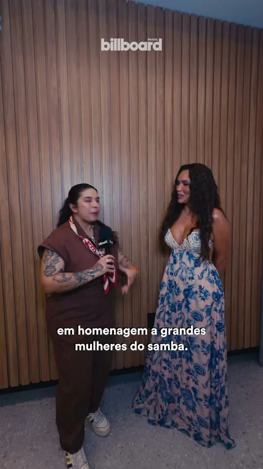 Karinah conversa com a entrevistadora da Billboard Brasil diante de uma parede de madeira