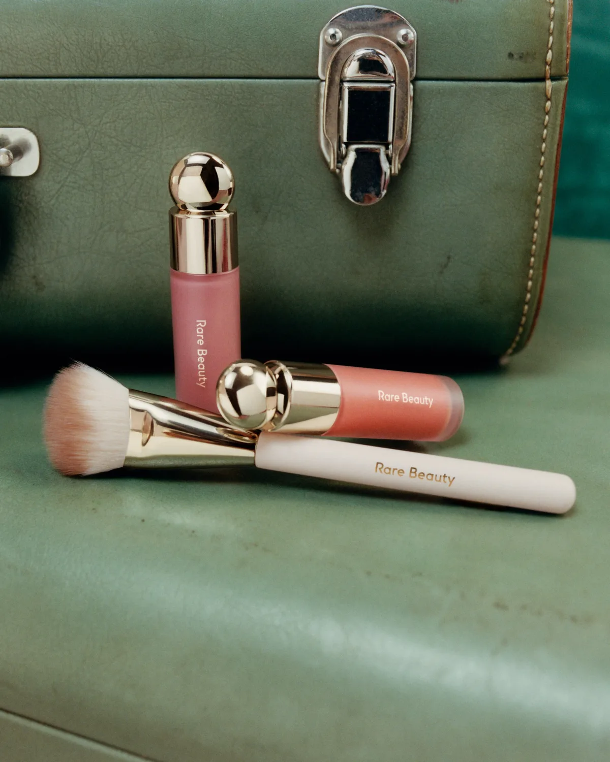 Blushes e pincel da coleção Delena sobre uma mala verde, na campanha da Rare Beauty