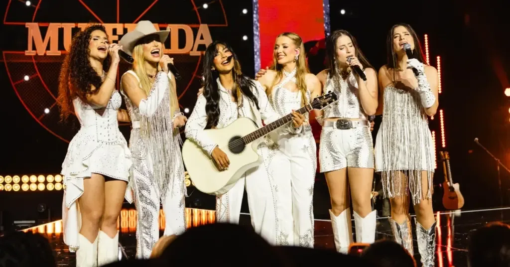 As seis integrantes da Muierada cantam juntas no palco, com figurinos claros.