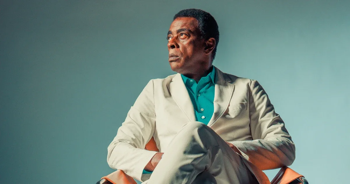 Seu Jorge sentado, com terno claro e camisa verde
